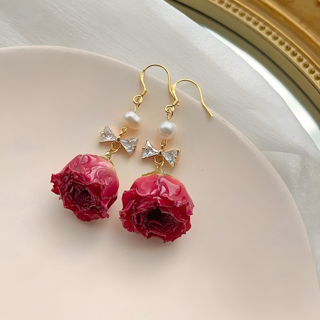 These are a pair of earrings with real flowers as the core design. They feature a unique craftsmanship: The delicate roses are preserved to freeze - frame their blooming posture. Matched with pearls, sparkling diamond - like bowknots and golden ear hooks, they blend natural romance with exquisite glamour, suitable for various styling occasions such as daily wear and dates. Wear flowers eternally and let the beauty of nature always accompany your ears.